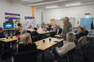 People attending a Launch North West business networking event at Leigh Spinners Mill, seated around tables while listening to a speaker in a bright meeting space.