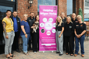 Group of volunteers and community partners standing together beside a Helping Wigan Thrive banner outside a community venue, smiling for a group photo.