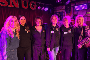 Launch North West team members and event volunteers standing together on stage at The Snug music venue during the Sound HER Out event, illuminated by purple stage lighting.