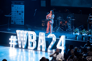 Bek Jones on stage at the Wigan Business Awards, with illuminated #WBA24 letters in the foreground, live entertainment setup behind, and guests seated in the audience.