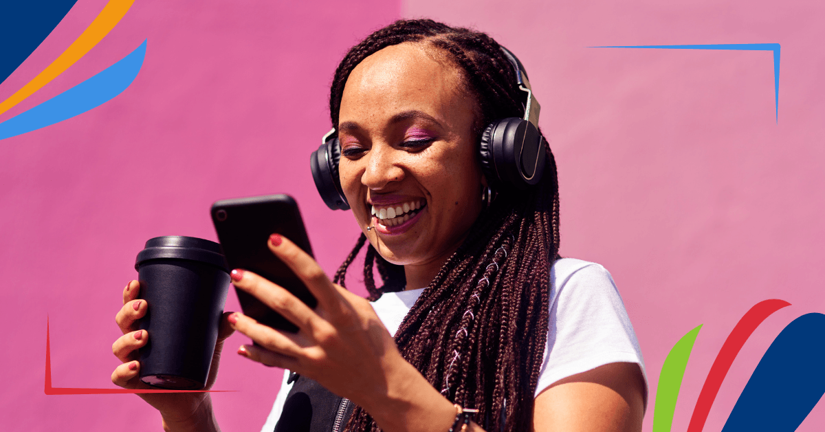 A person wearing headphones and holding a coffee cup looks at a smartphone while smiling against a pink background.